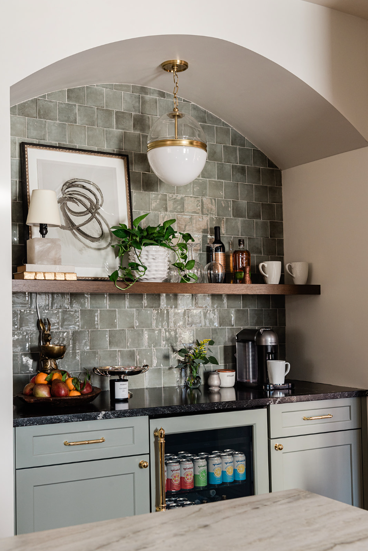 Styled home bar with pale green cabinets, a stone counter and an open wood shelf.